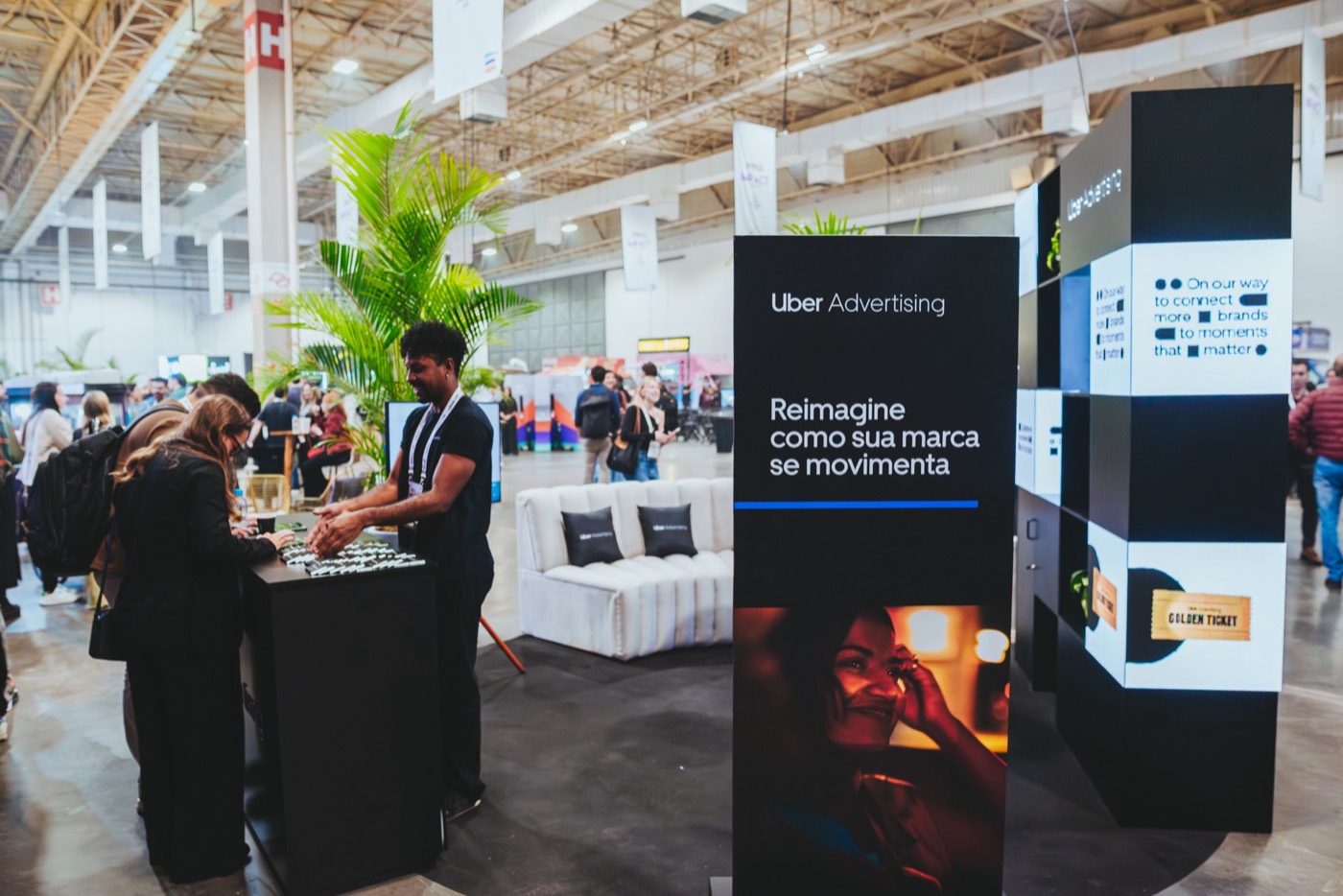 Black pillar reading "Uber Advertising" and "Reimagine como sua marca se movimenta" at a booth, people at a high table