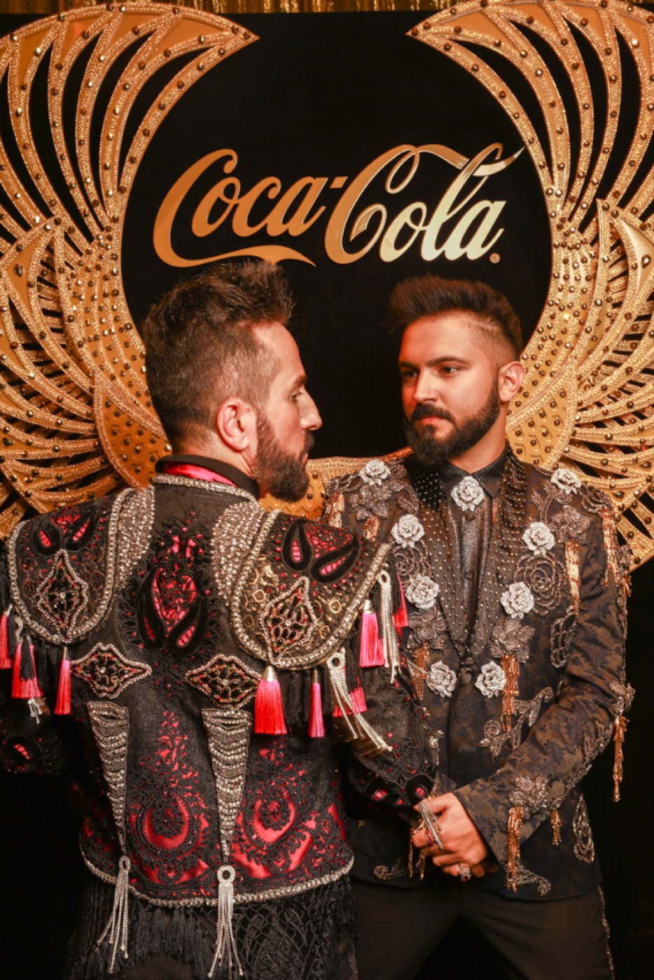 Two people in embroidered jackets look at each other before a backdrop with golden wings and a "Coca-Cola" logo