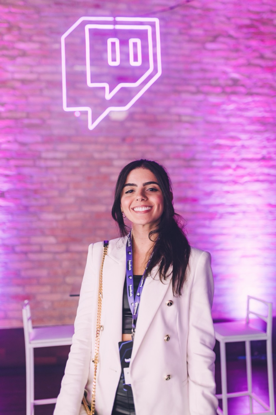 Person in a white blazer with a badge smiles in front of a brick wall with a neon logo