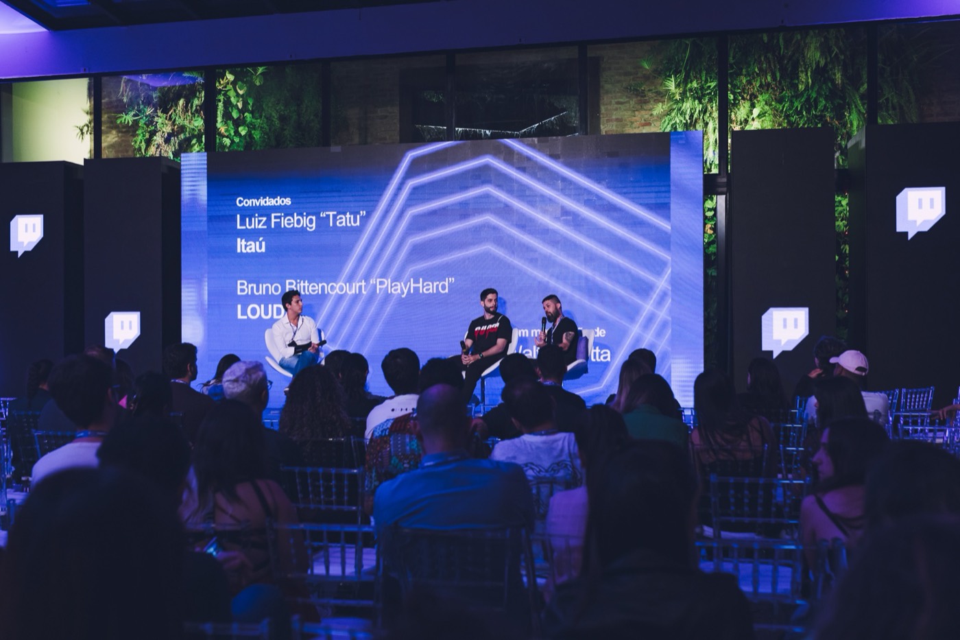 Three people talk on stage in front of a blue screen while the audience watches seated