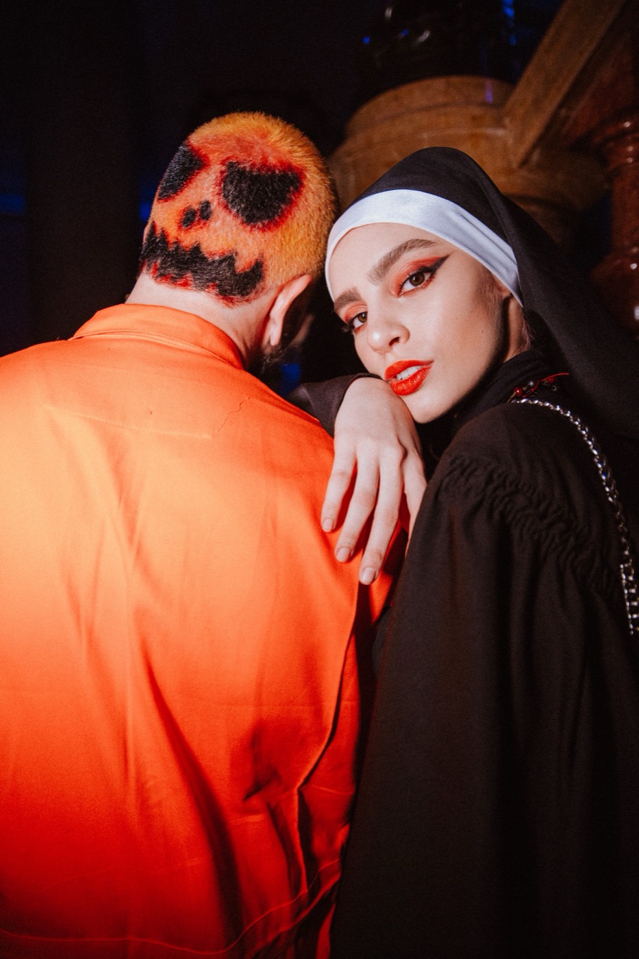 Person dressed as a nun rests a hand on someone seen from behind, a pumpkin face drawn in their hair