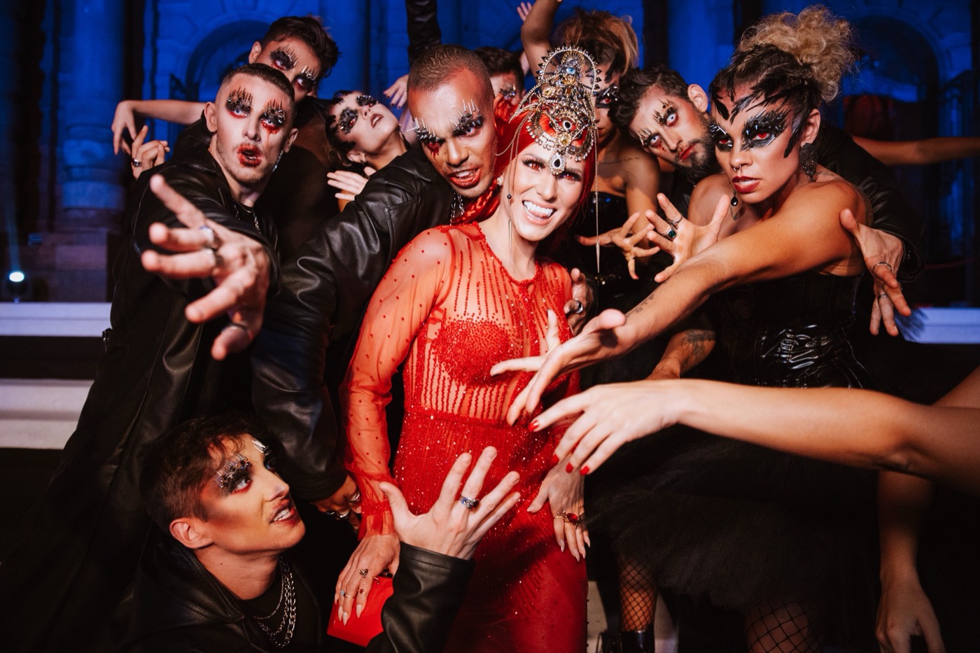 Person in a red dress and headpiece poses surrounded by a group in dark makeup with outstretched hands