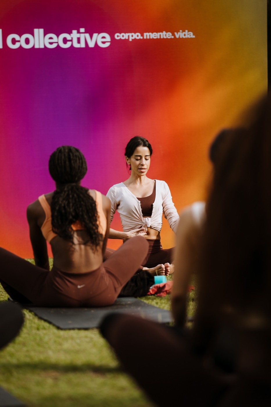Two people seated facing each other on a mat on the grass, before a colorful panel with the word "collective"