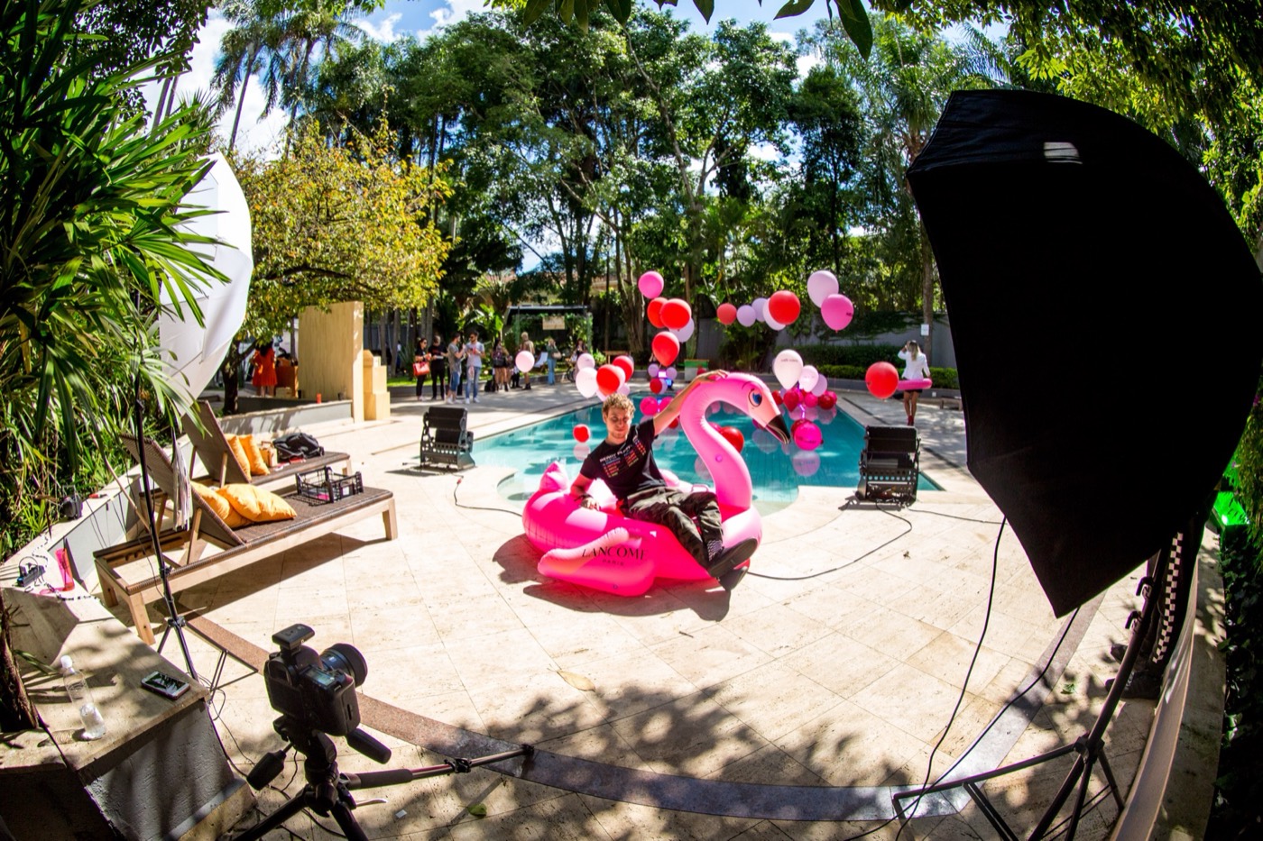 Poolside photo set with a camera on a tripod, lighting gear and a person seated on a pink flamingo float