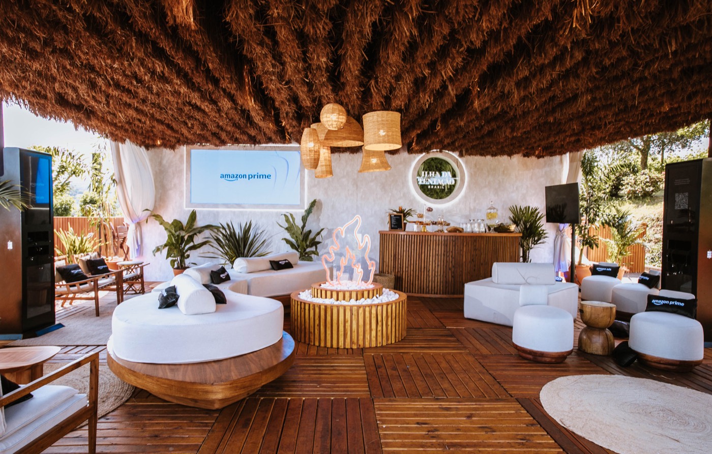 Lounge under a thatched roof with round white sofas, a central neon campfire, a bar and a screen behind