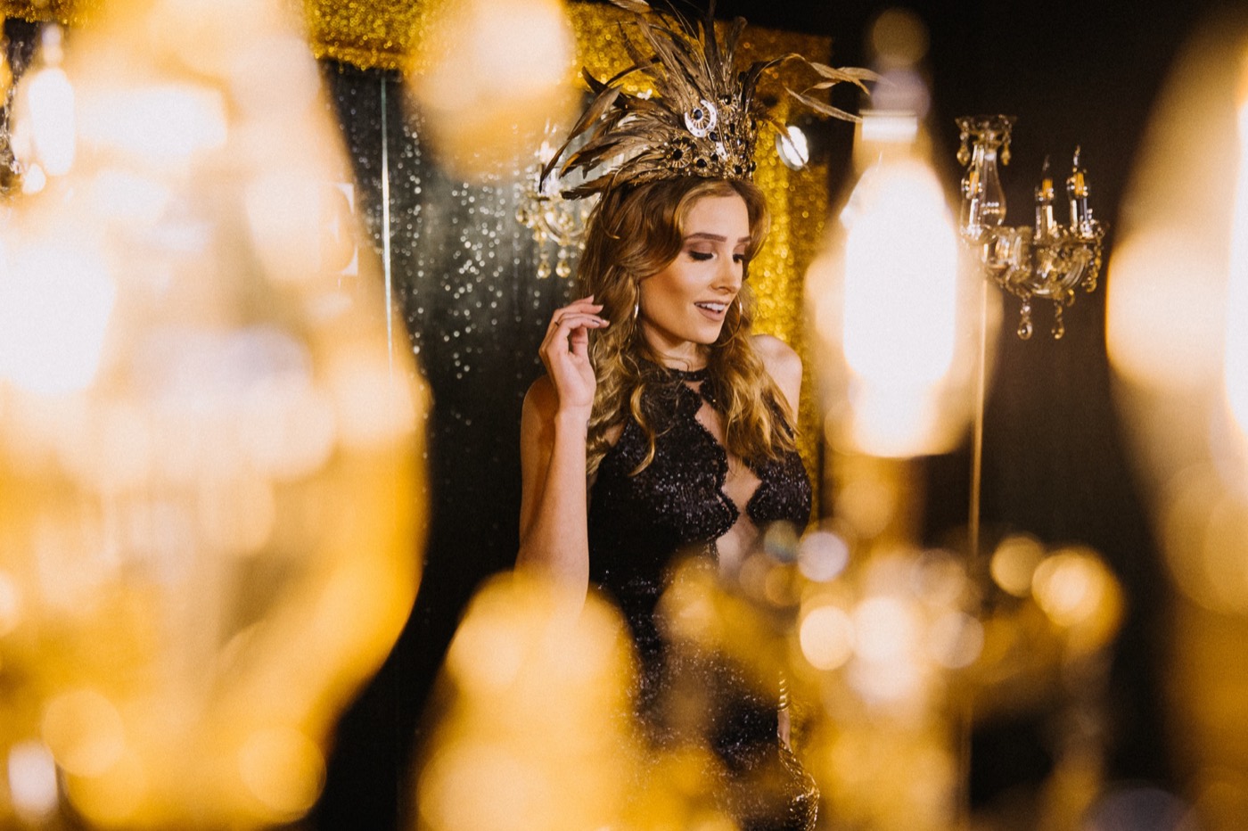 Person in a feathered headpiece and sparkling black dress, seen through blurred golden chandeliers