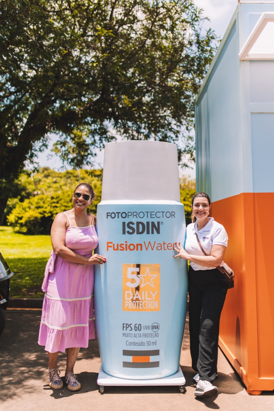 Two people pose smiling beside a giant "ISDIN Fusion Water" bottle replica outdoors
