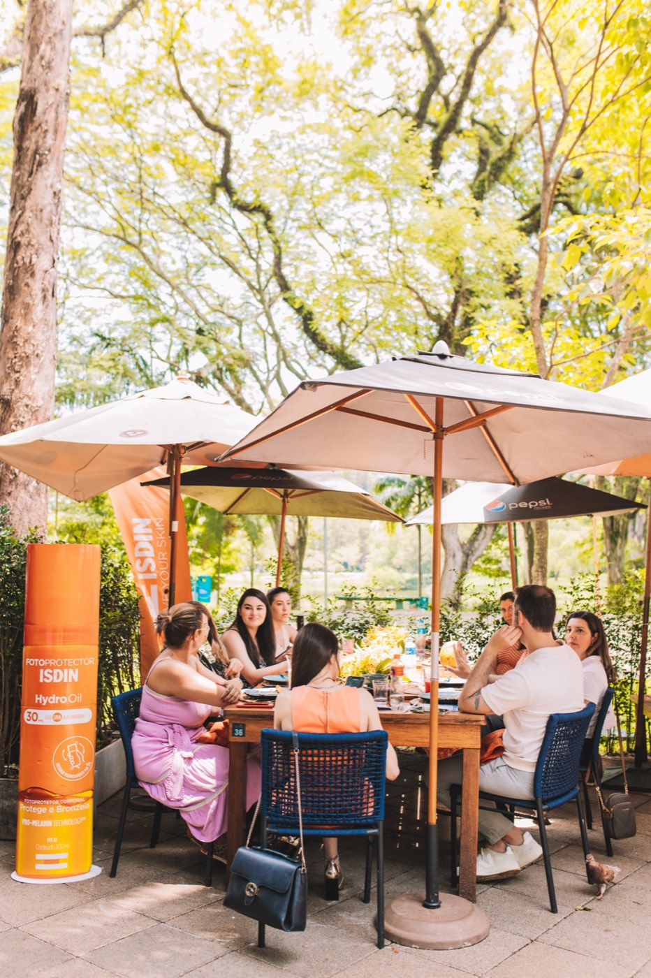 Group chats at an outdoor table under umbrellas and trees, beside an orange bottle-shaped totem