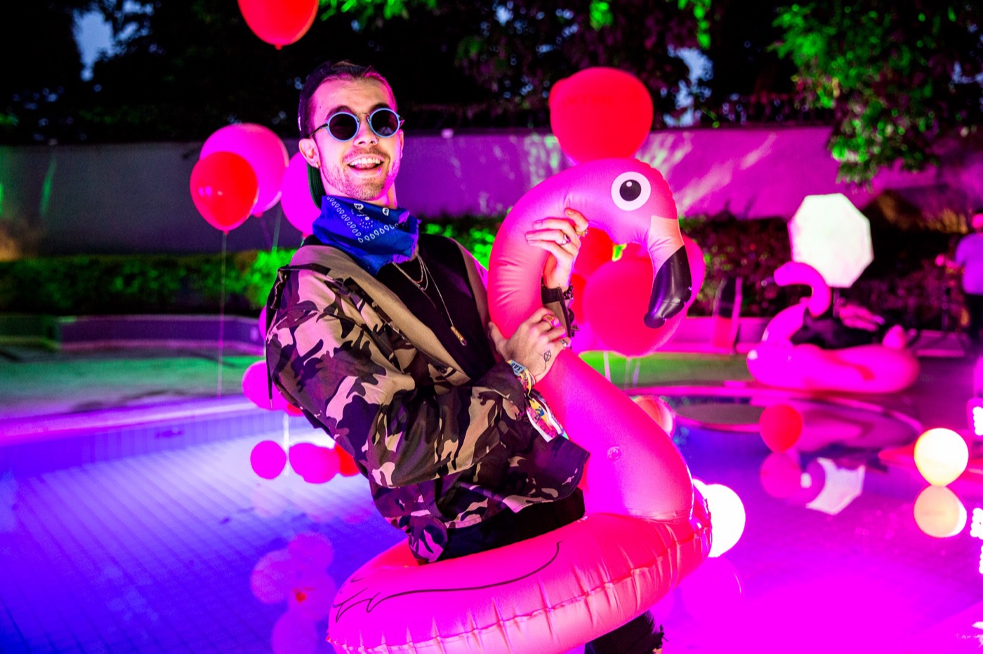 Person in sunglasses and a camouflage jacket poses inside a flamingo float in a pool with pink and purple light
