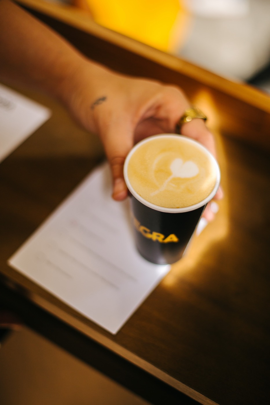 Hand holds a black coffee cup with a heart drawn in the foam, seen from above over a counter