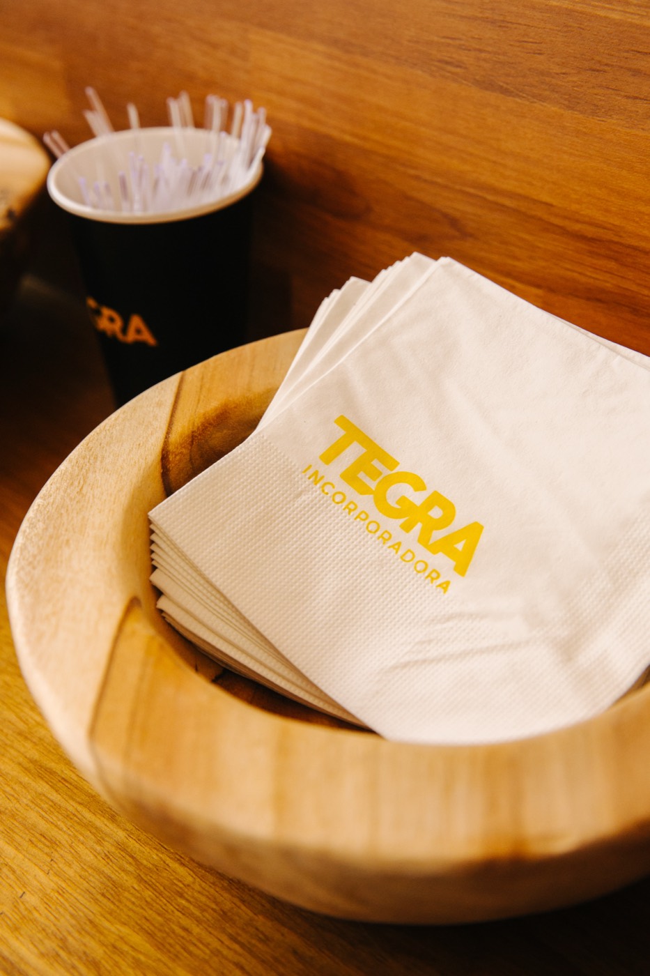 White napkins printed with "TEGRA INCORPORADORA" in yellow in a wooden bowl, next to a cup of stirrers