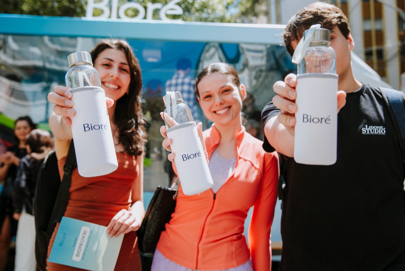 Three people smile and hold out glass bottles with white "Bioré" sleeves toward the camera
