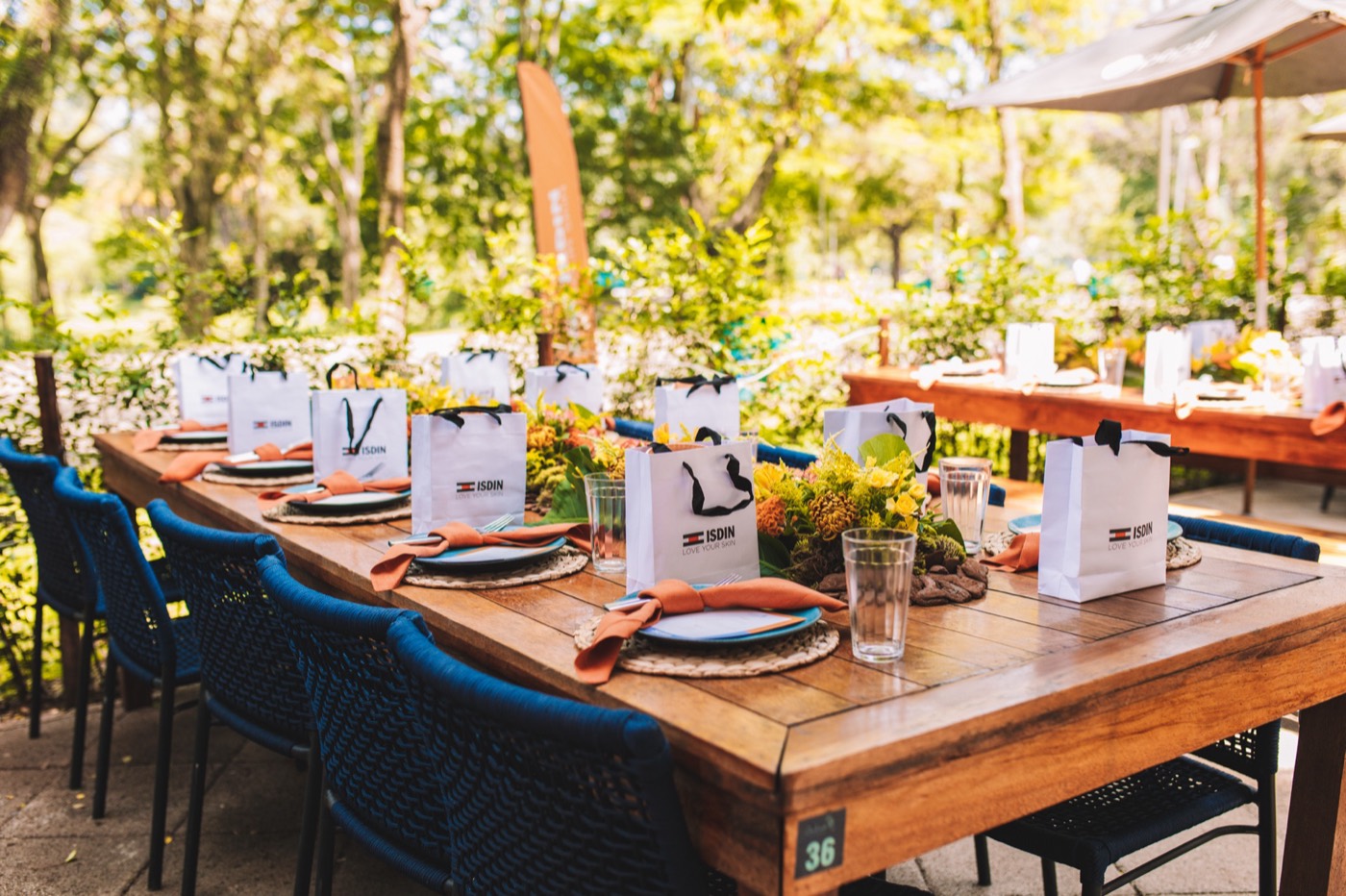 Long wooden outdoor table set with plates, orange napkins, flowers and white "ISDIN" bags
