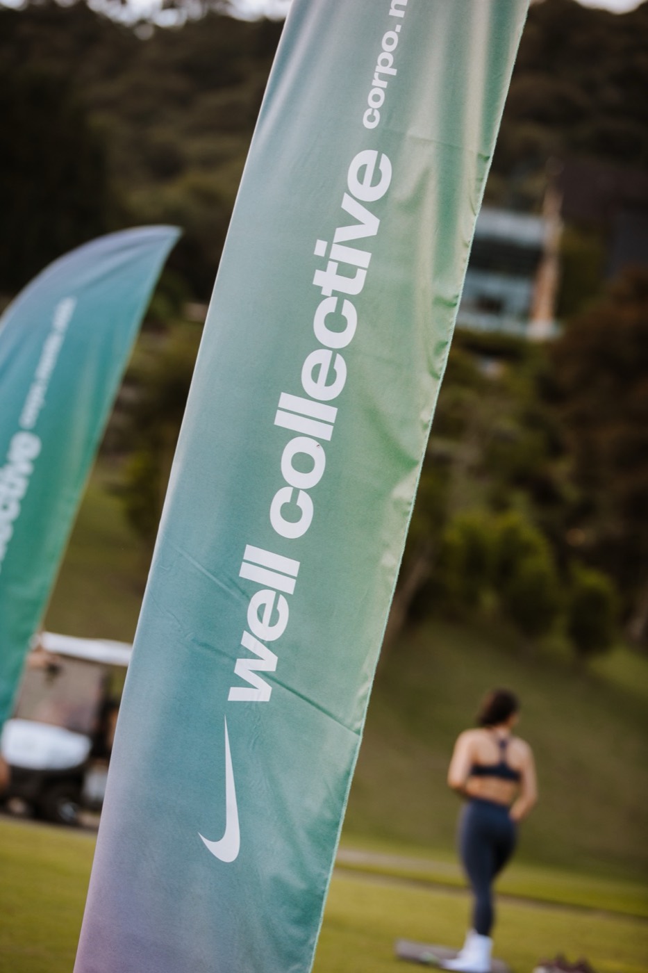 Vertical green-toned flags reading "well collective" on a lawn, with a person seen from behind in the background