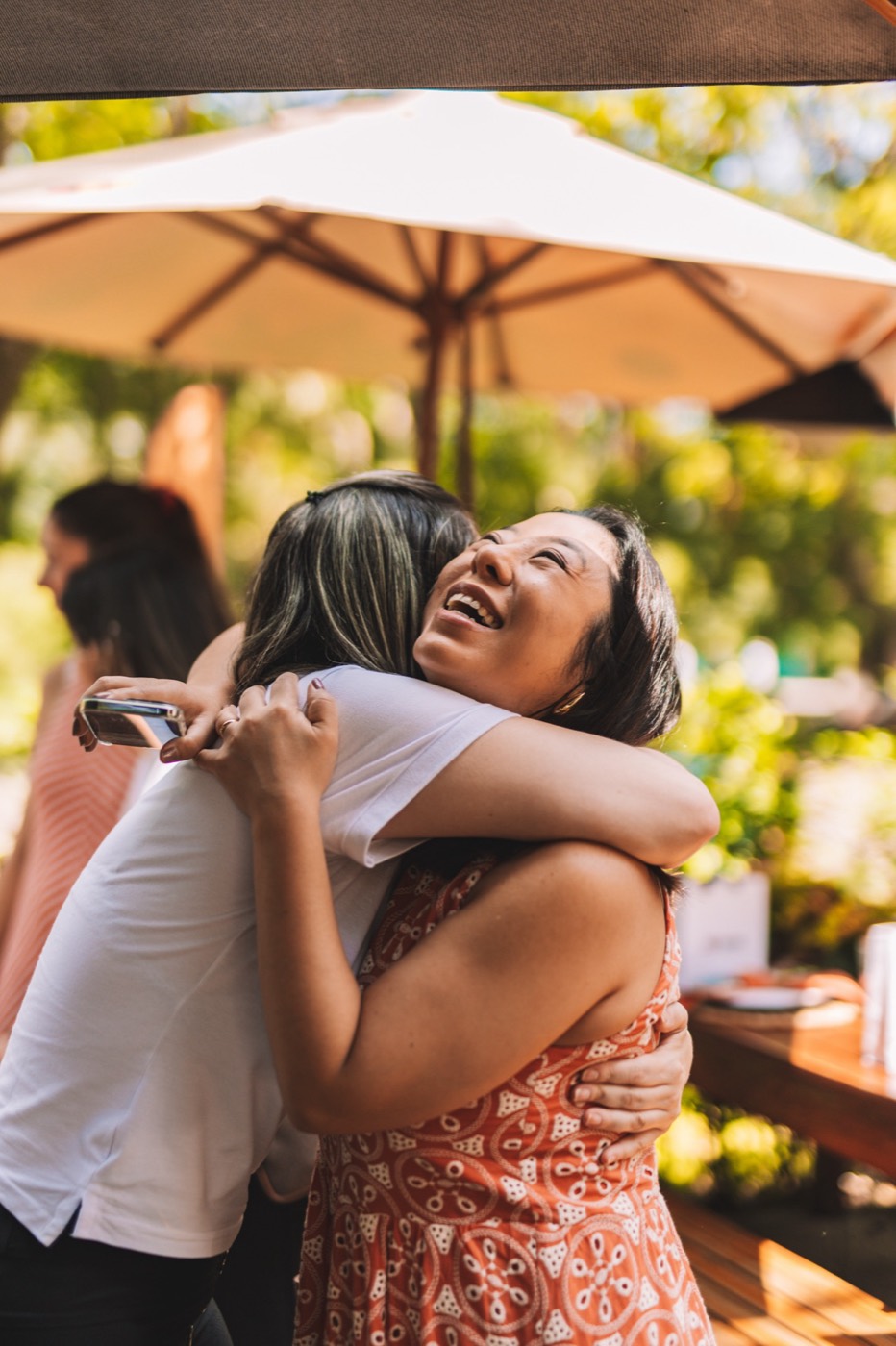 Two people hug smiling under an umbrella in a leafy outdoor area, one of them holding a phone