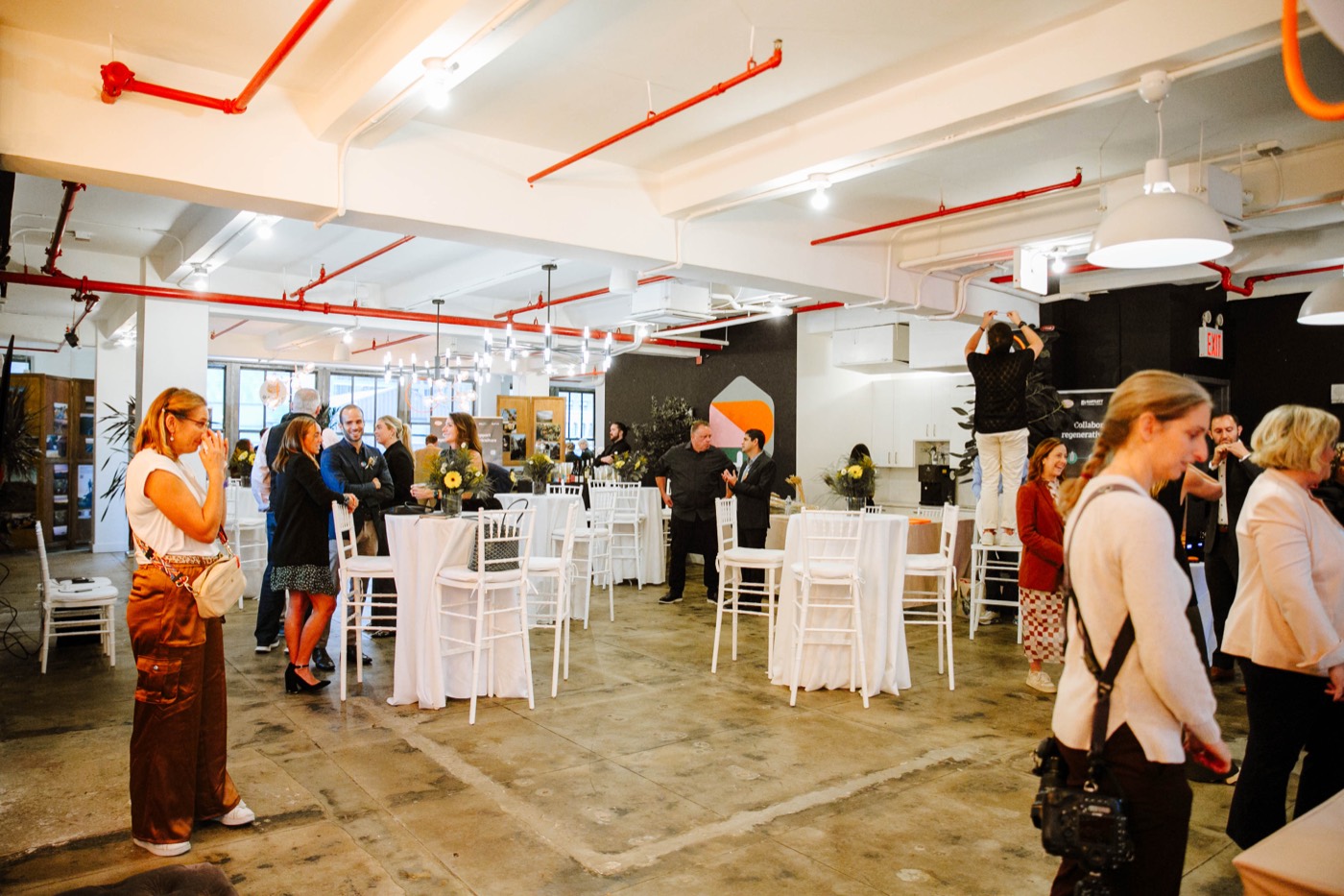 Guests chat standing around white high tables in a spacious hall with red pipes on the ceiling