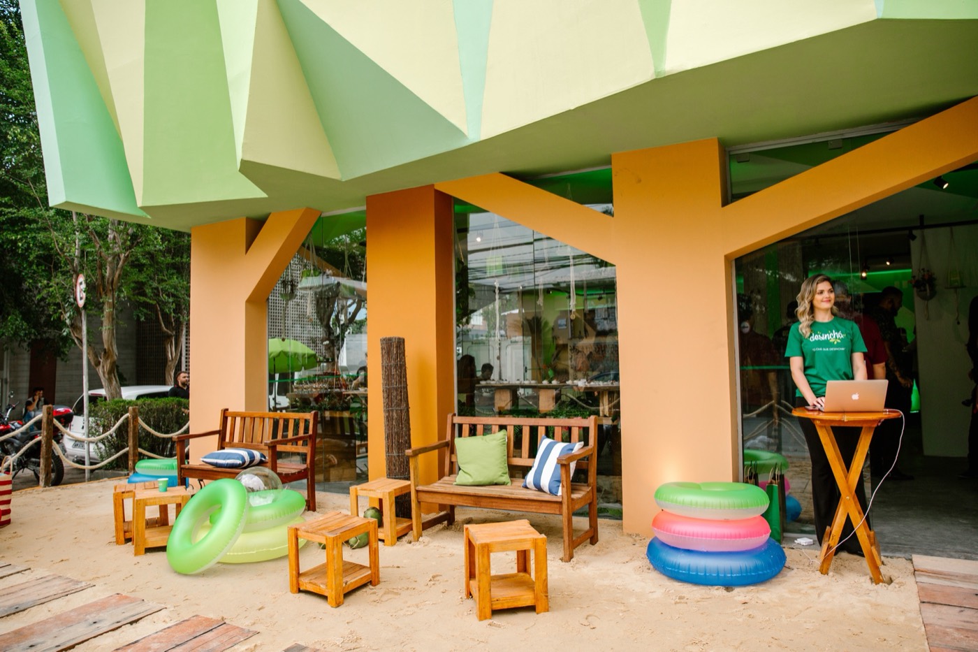 Outdoor area with sand, wooden benches, colorful pool floats and a person in a green T-shirt at a laptop stand
