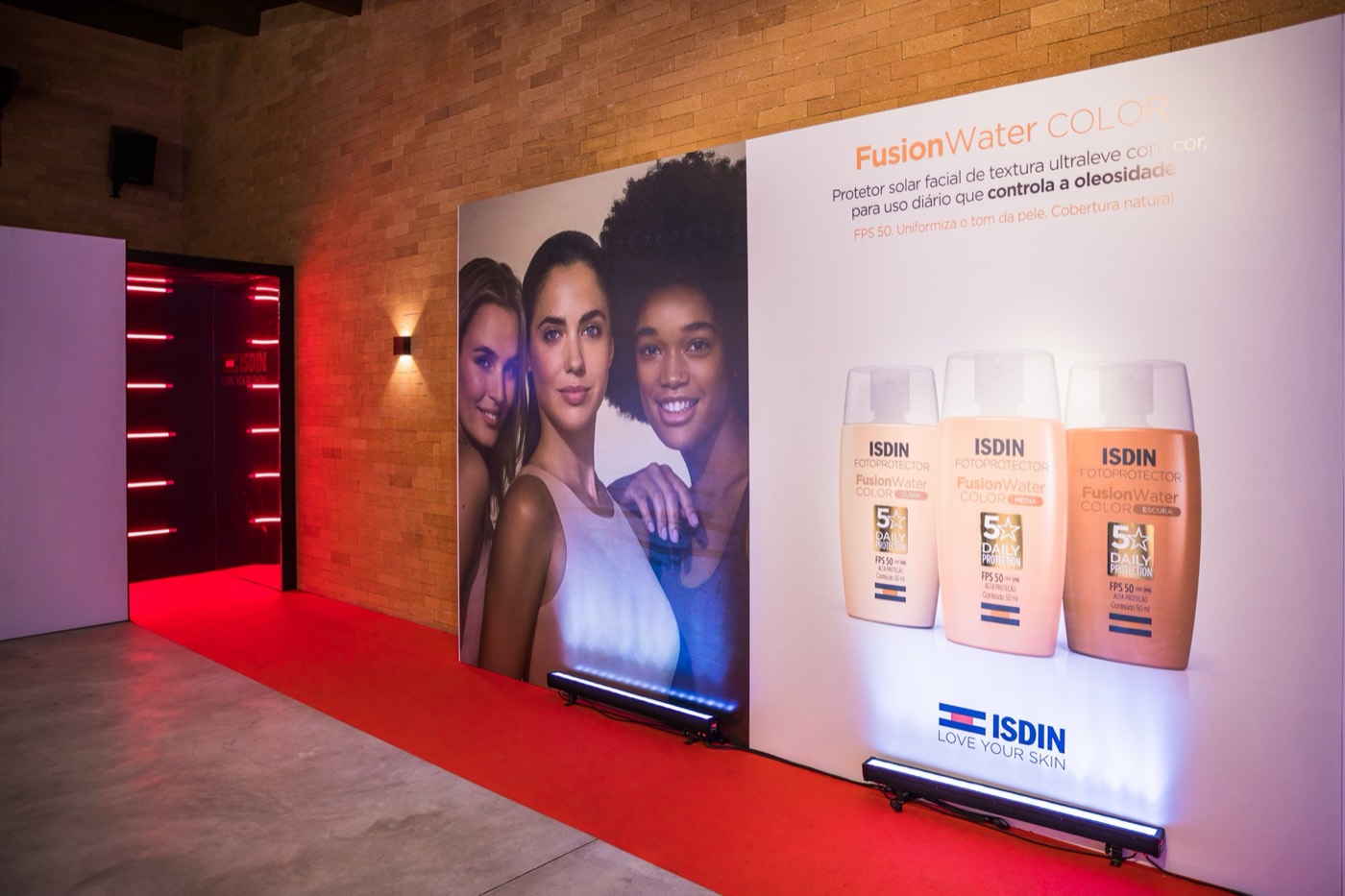 Printed panel with three faces and three "ISDIN" sunscreen bottles beside a corridor with a red floor