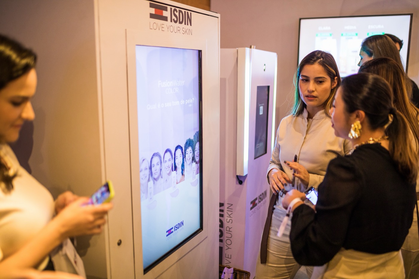 People in front of white kiosks with vertical screens and the "ISDIN" brand, one of them using a phone