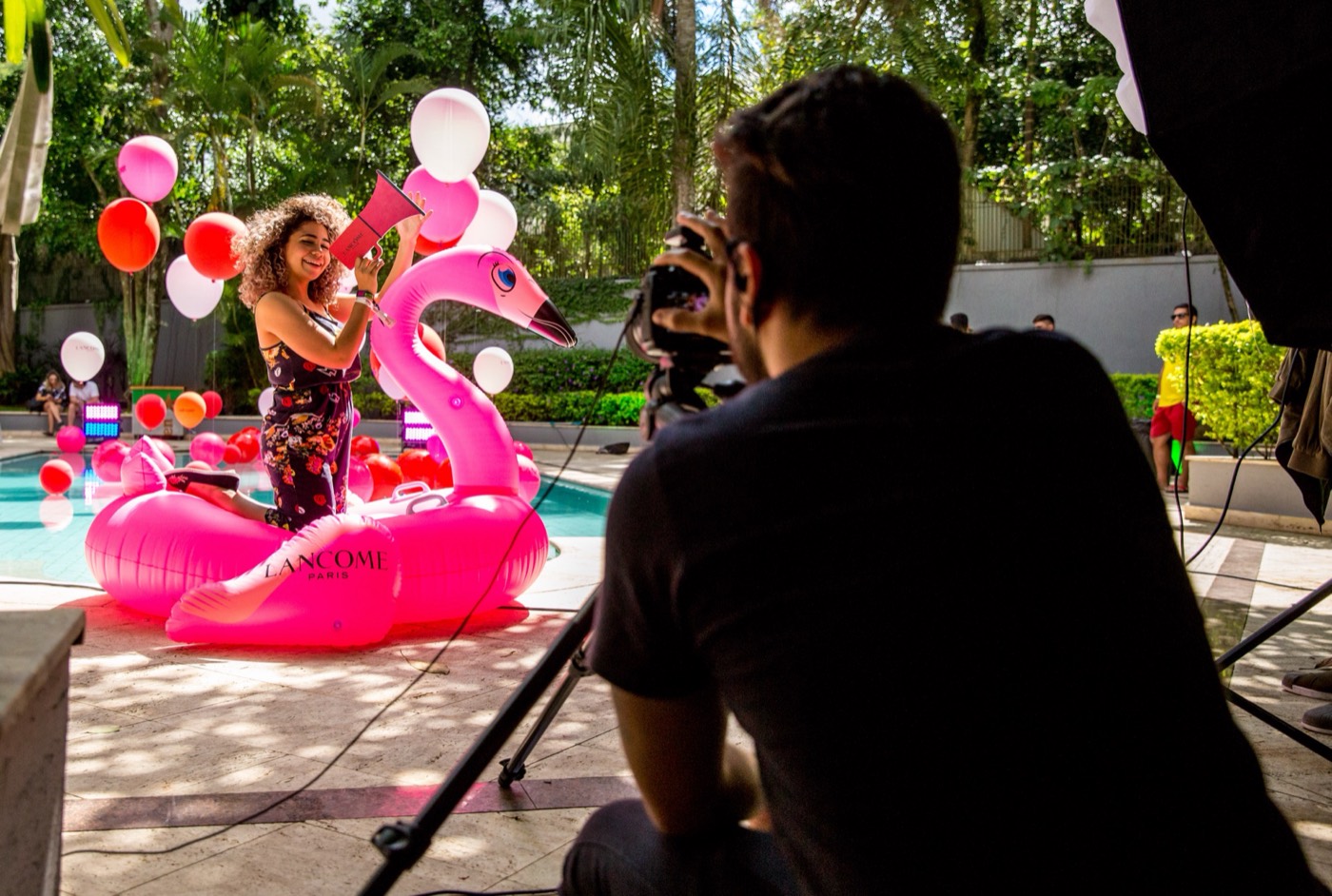 Person seen from behind photographs another holding a pink megaphone on a "LANCÔME" flamingo float by the pool
