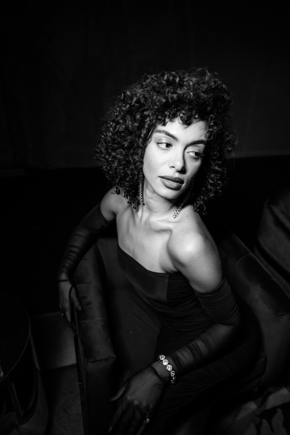 Black and white portrait of a person with curly hair, a dark dress and gloves, seated in an armchair