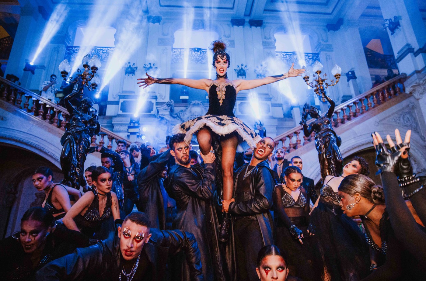 Person lifted with arms outstretched by a group dressed in black, in front of a staircase lit in blue