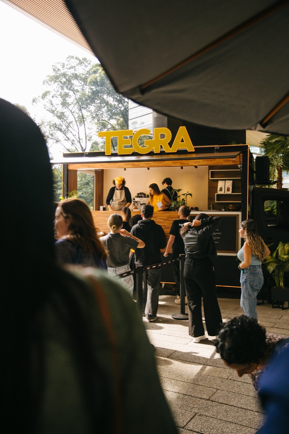 Coffee trailer with a "TEGRA" sign and a line of people, seen from behind someone blurred in the foreground