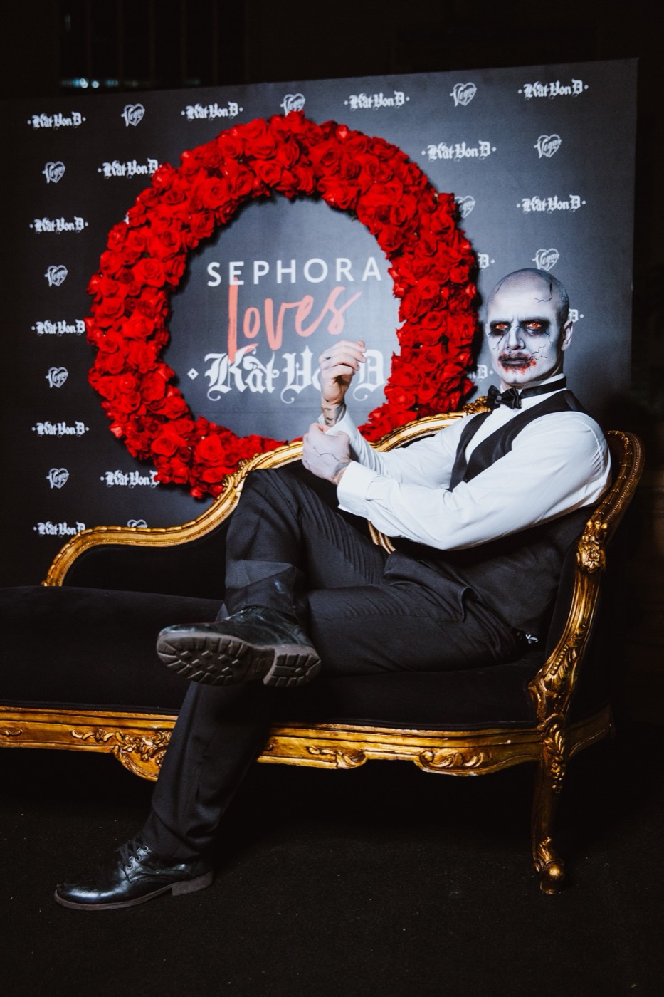 Person with a white-painted face on a gold sofa, before a backdrop with a red rose wreath and "SEPHORA Loves"