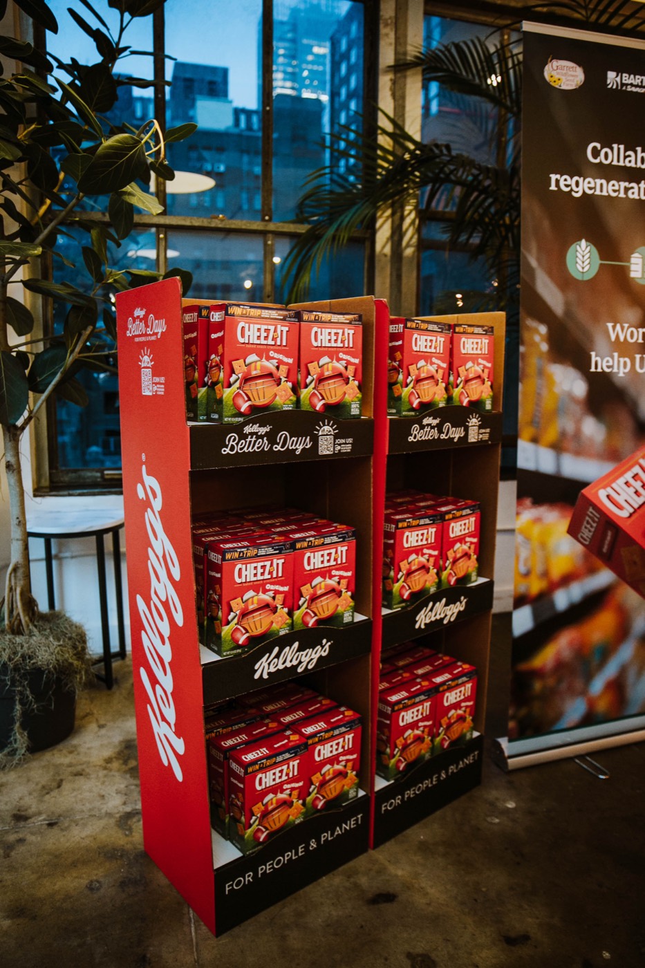 Red three-shelf display with "CHEEZ-IT" boxes and the "Kellogg's" brand by a window at dusk