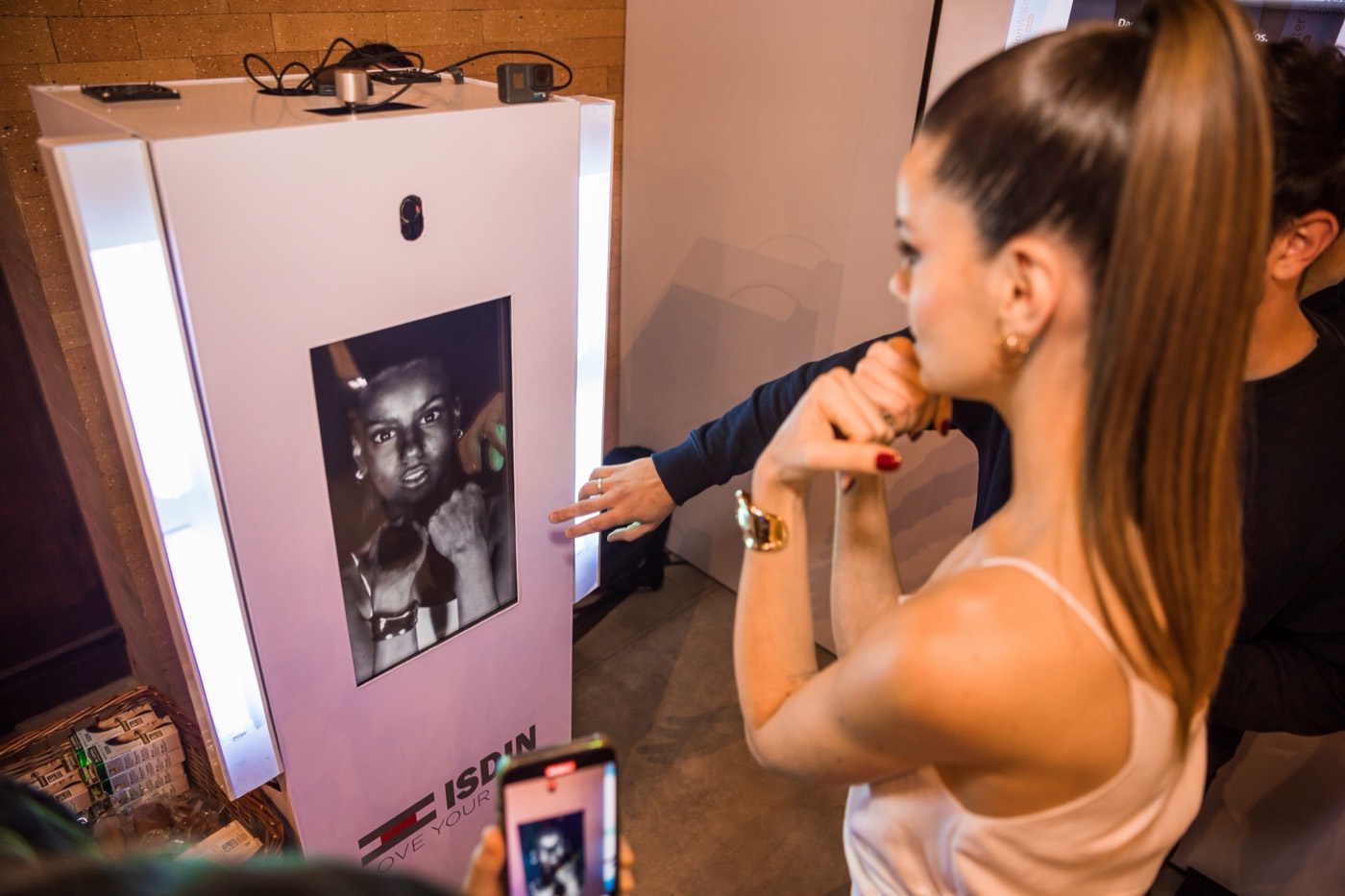 Person with a ponytail poses in front of a white photo booth displaying a black and white portrait