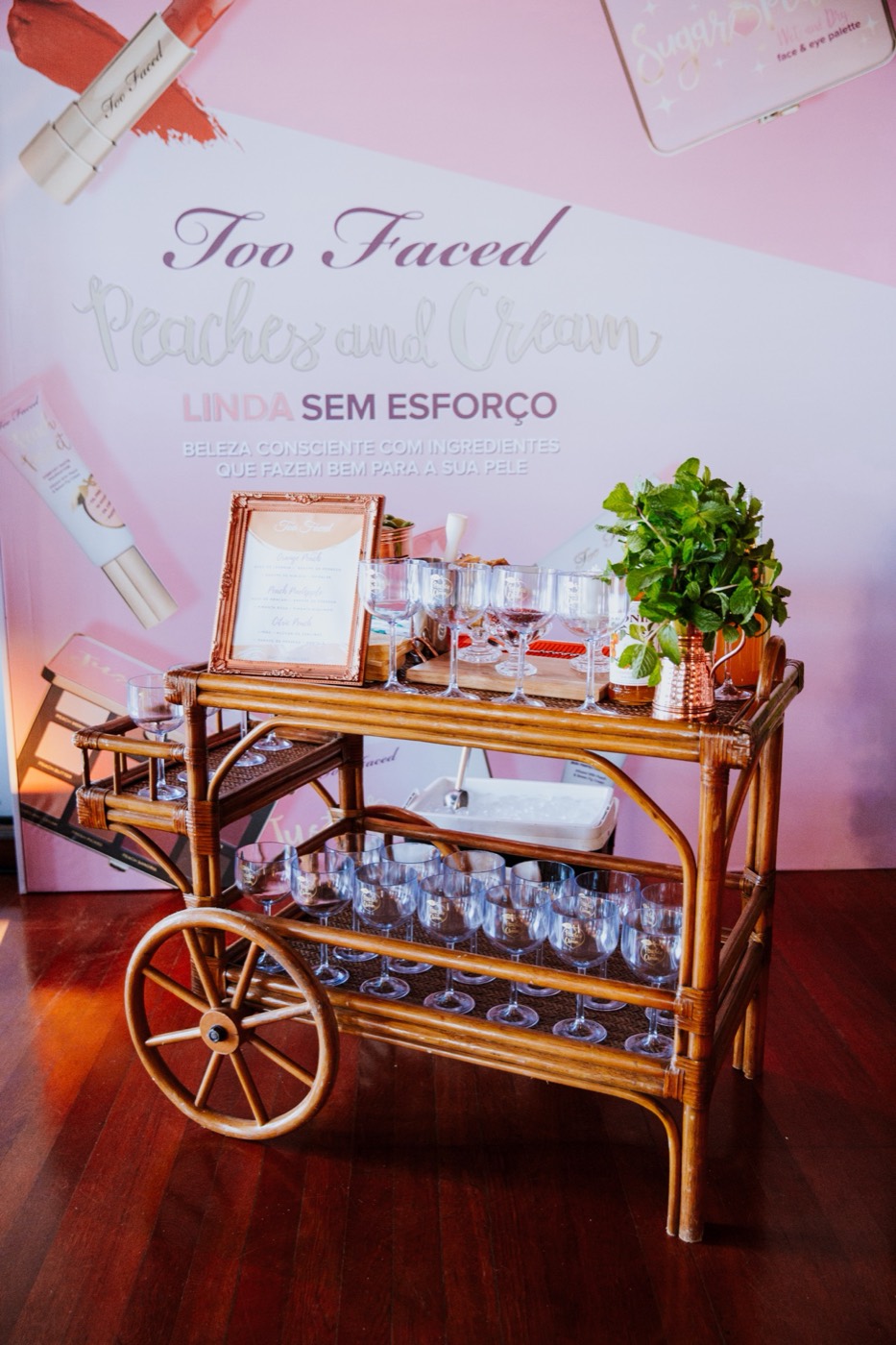 Wicker bar cart with glasses and a potted plant in front of a pink "Too Faced Peaches and Cream" panel