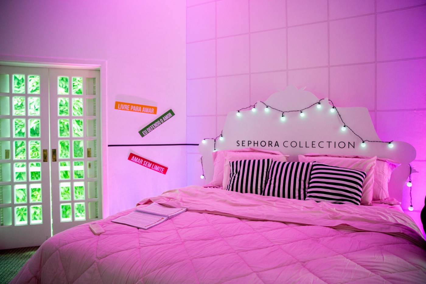 Staged bedroom in pink light, a bed with striped pillows and a lit headboard reading "SEPHORA COLLECTION"