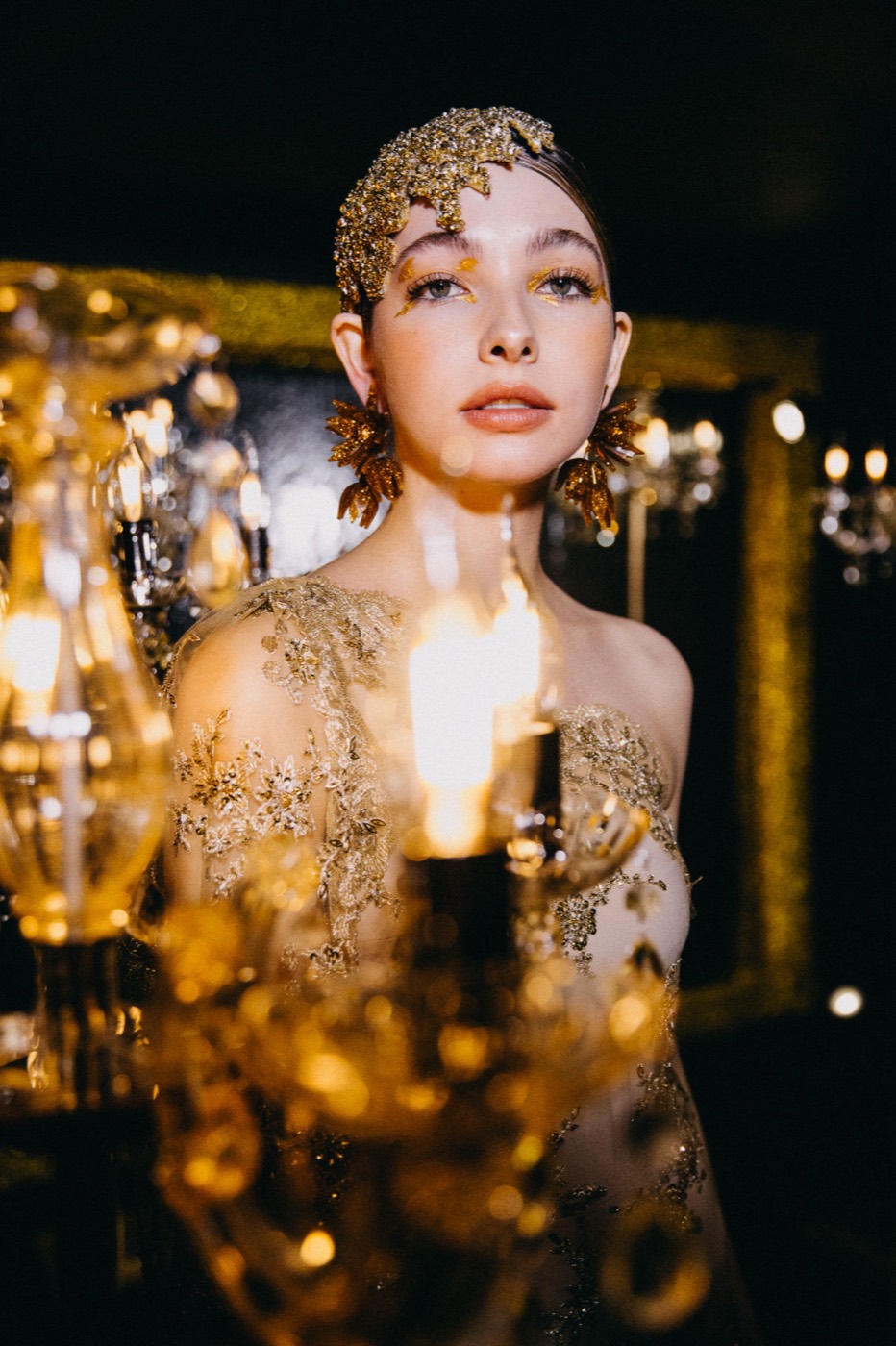 Person in a gold headpiece and embroidered dress, behind a lit crystal chandelier