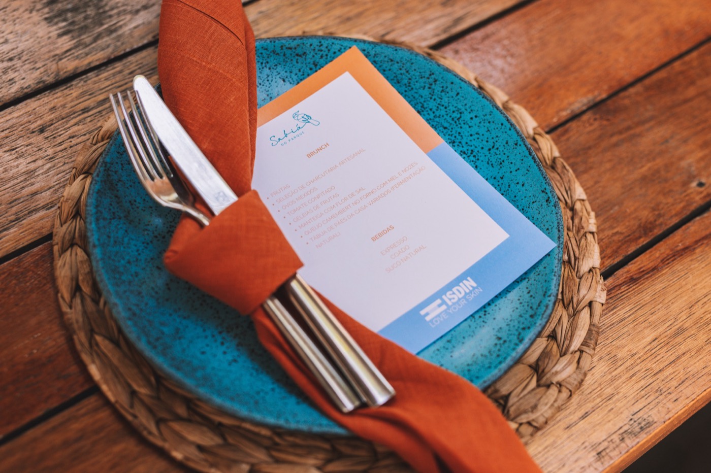 Blue plate on a straw placemat, with cutlery in an orange napkin and a printed menu bearing the "ISDIN" brand