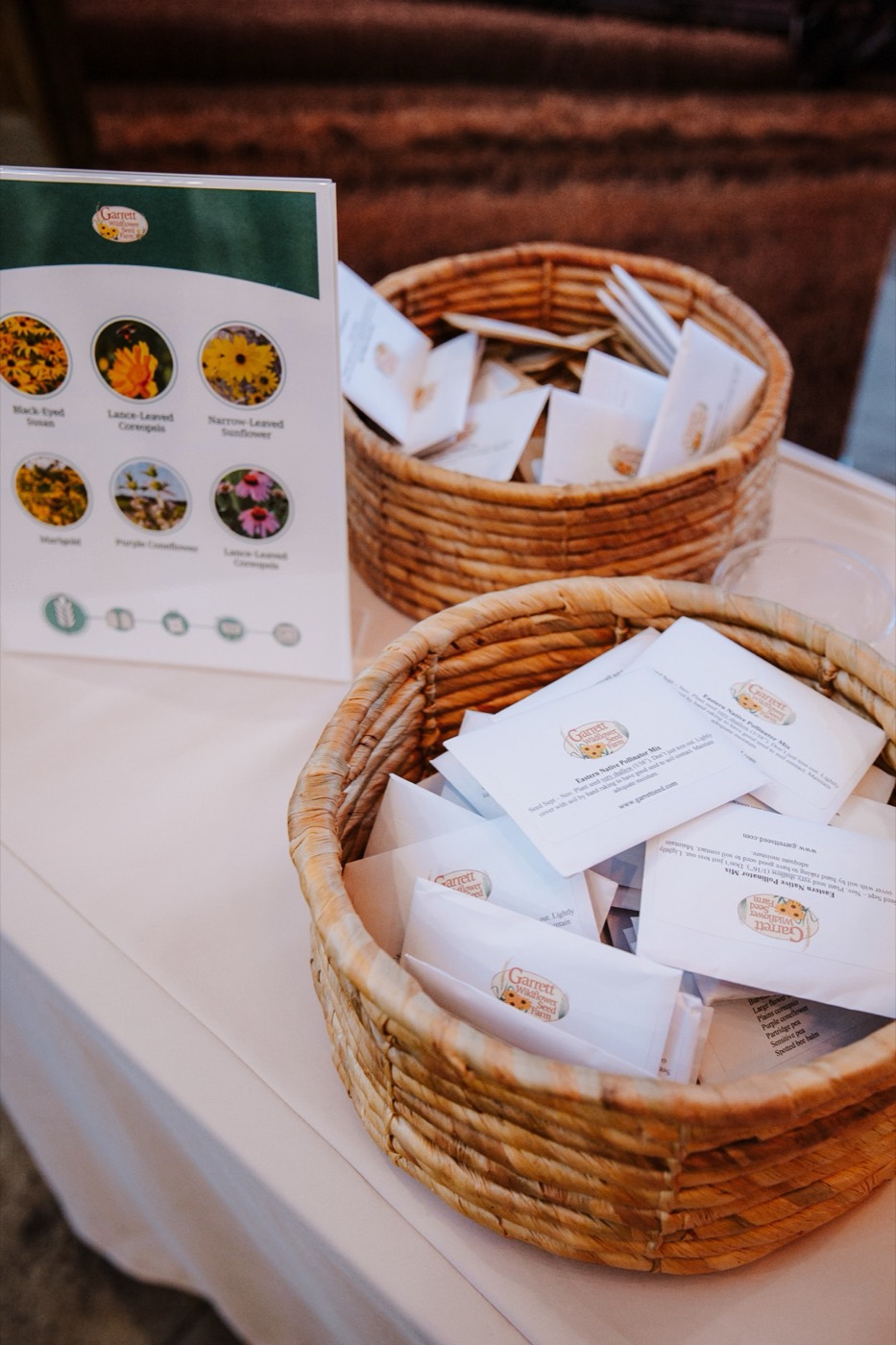 Straw baskets with printed white envelopes on a table, beside a sign with photos of flowers
