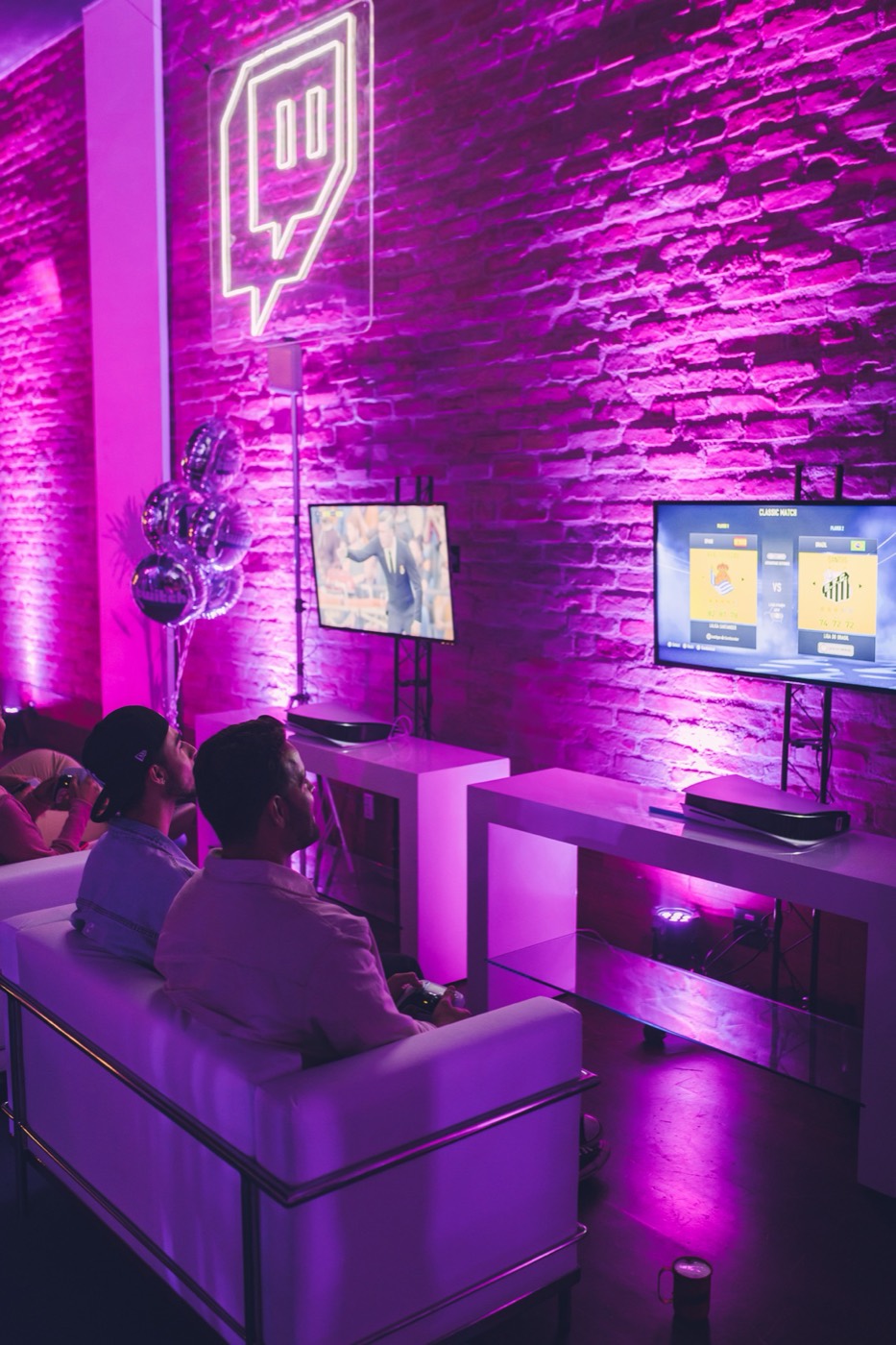 Side view of gaming stations with two screens, metallic balloons and people seated on a white sofa