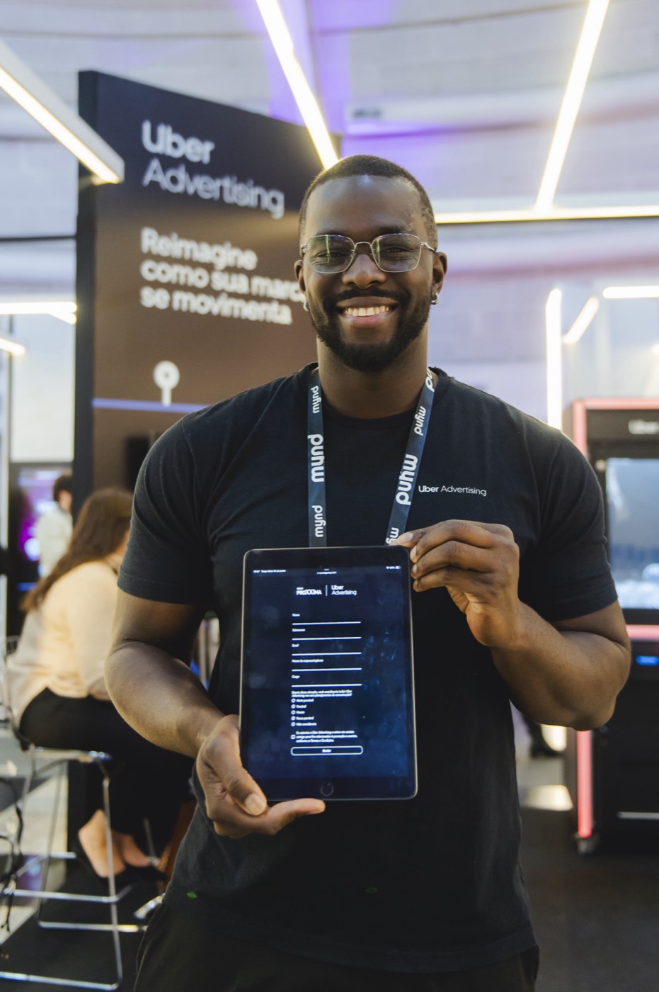 Smiling person in a black T-shirt shows a tablet with a form on screen, before an "Uber Advertising" panel
