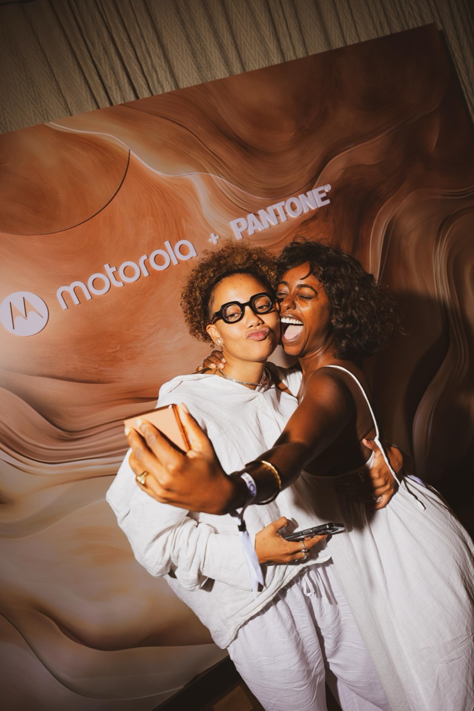 Two people hug and take a selfie in front of a brown-toned panel reading "motorola + PANTONE"