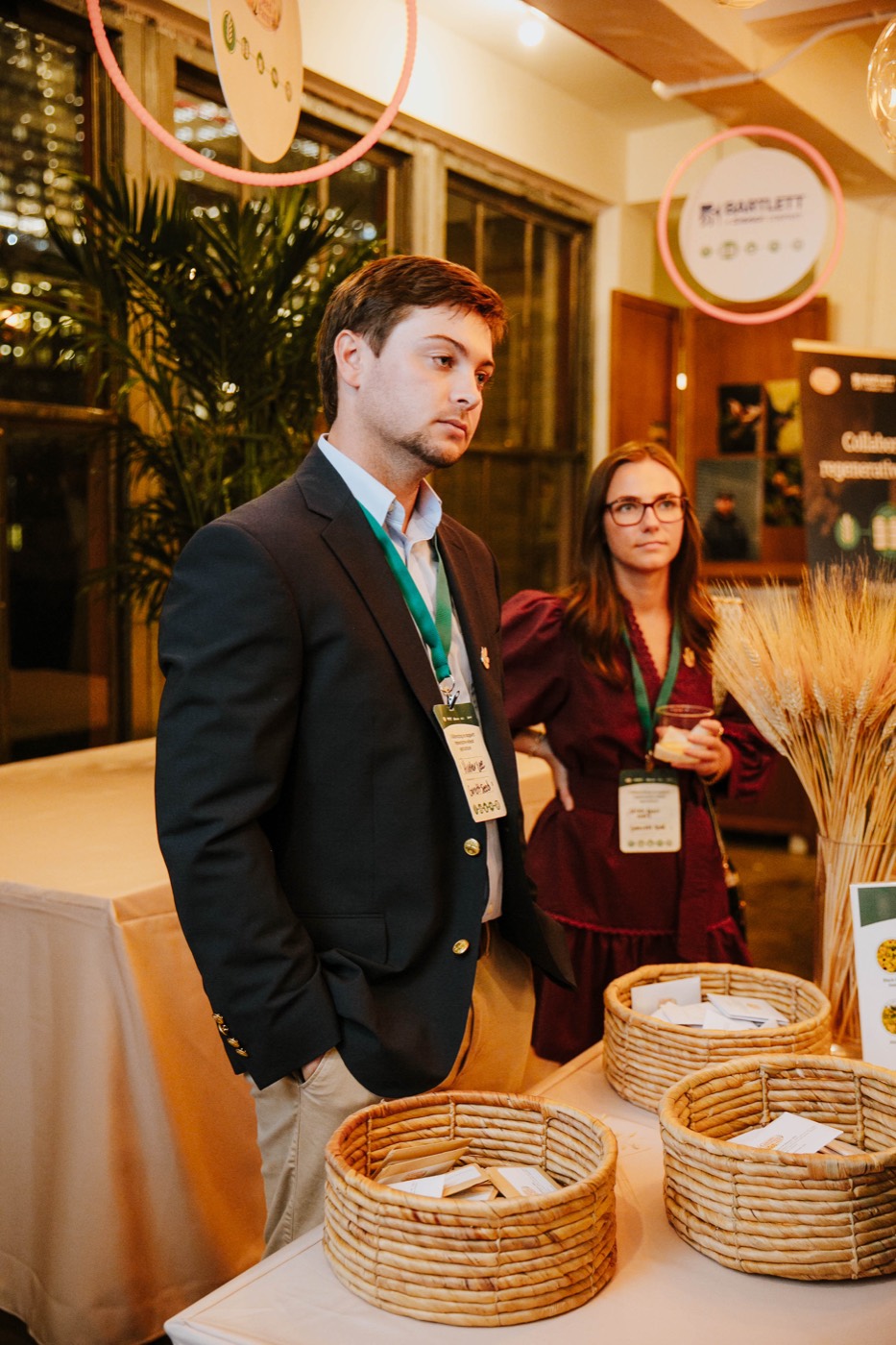 Two people wearing badges behind a table with straw baskets, beside an arrangement of wheat stalks