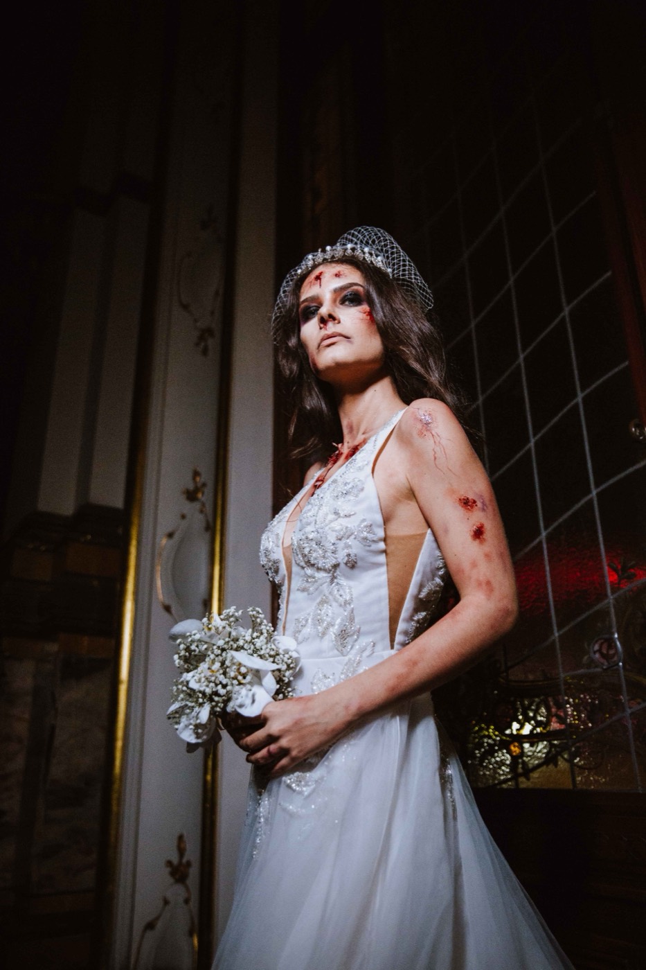 Person dressed as a bride in a white gown, veil and bouquet, with red stains simulating blood
