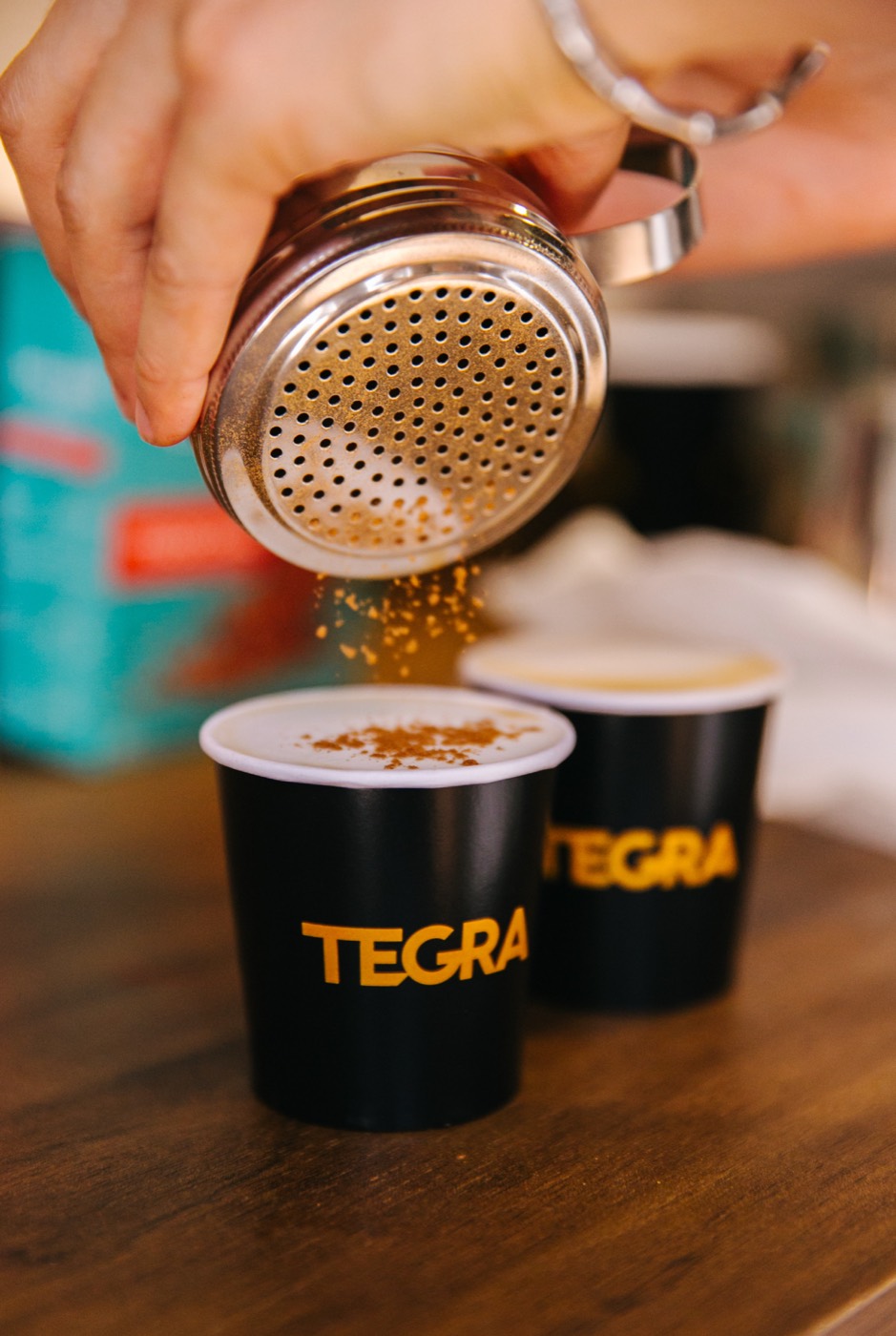 Hand uses a metal shaker over the coffee foam in black cups reading "TEGRA"