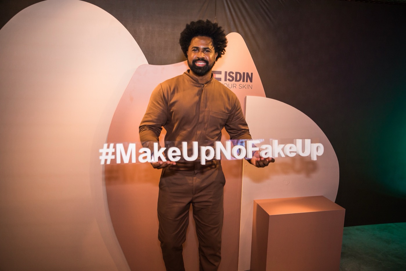 Smiling person poses in front of an earth-toned backdrop with the hashtag "#MakeUpNoFakeUp" in white letters