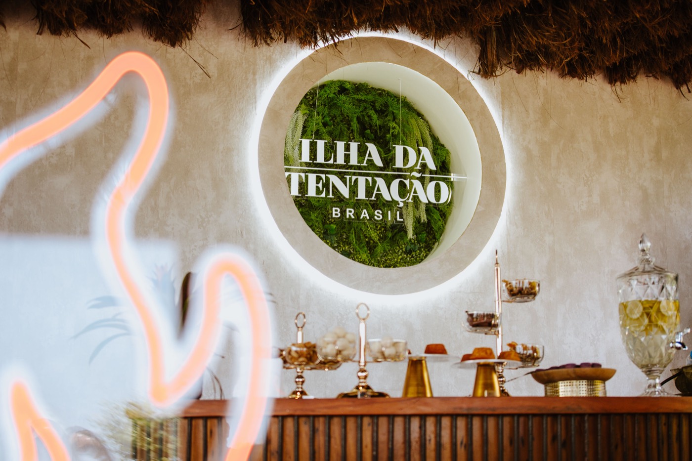 Round backlit "ILHA DA TENTAÇÃO BRASIL" sign on a foliage background, above a bar with sweets and a drink dispenser