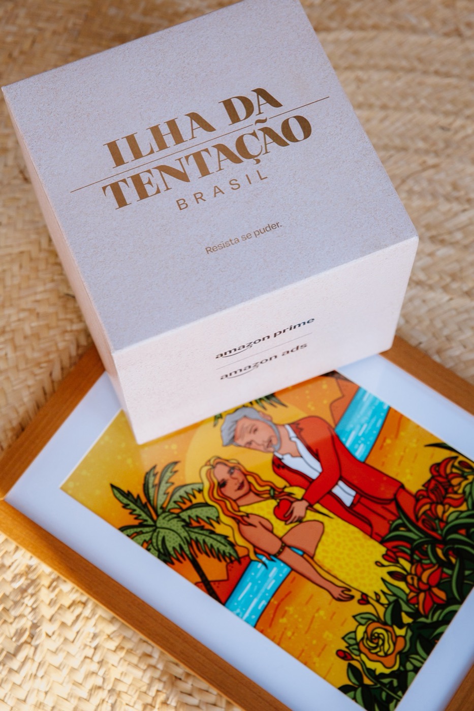 White box with "ILHA DA TENTAÇÃO BRASIL" in gold on a framed colorful illustration of a couple in tropical scenery