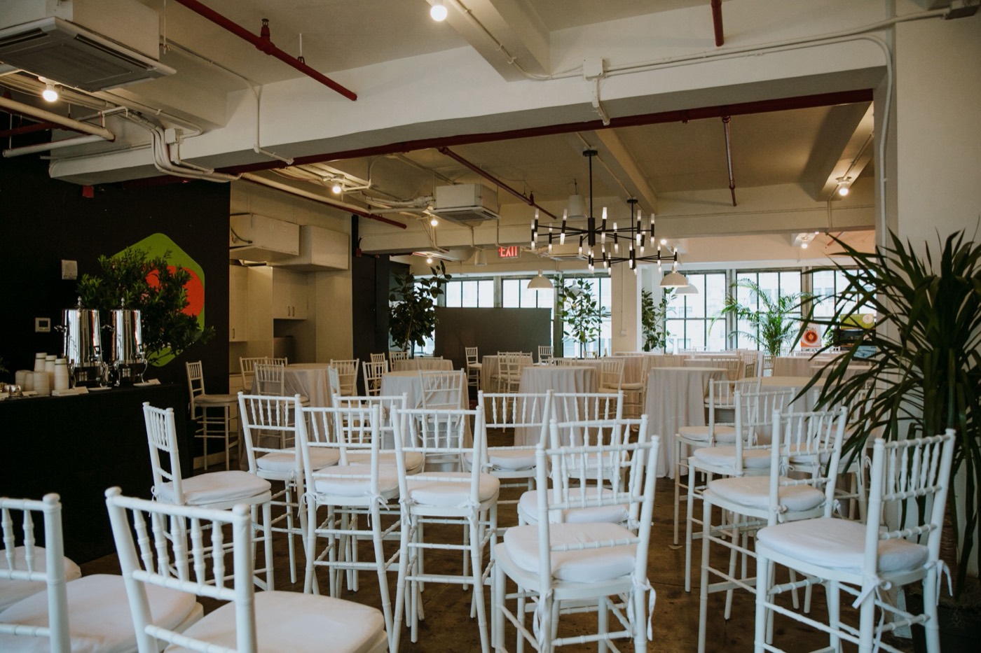 Rows of white chairs in a hall with a chandelier, plants and a coffee station with urns to the side