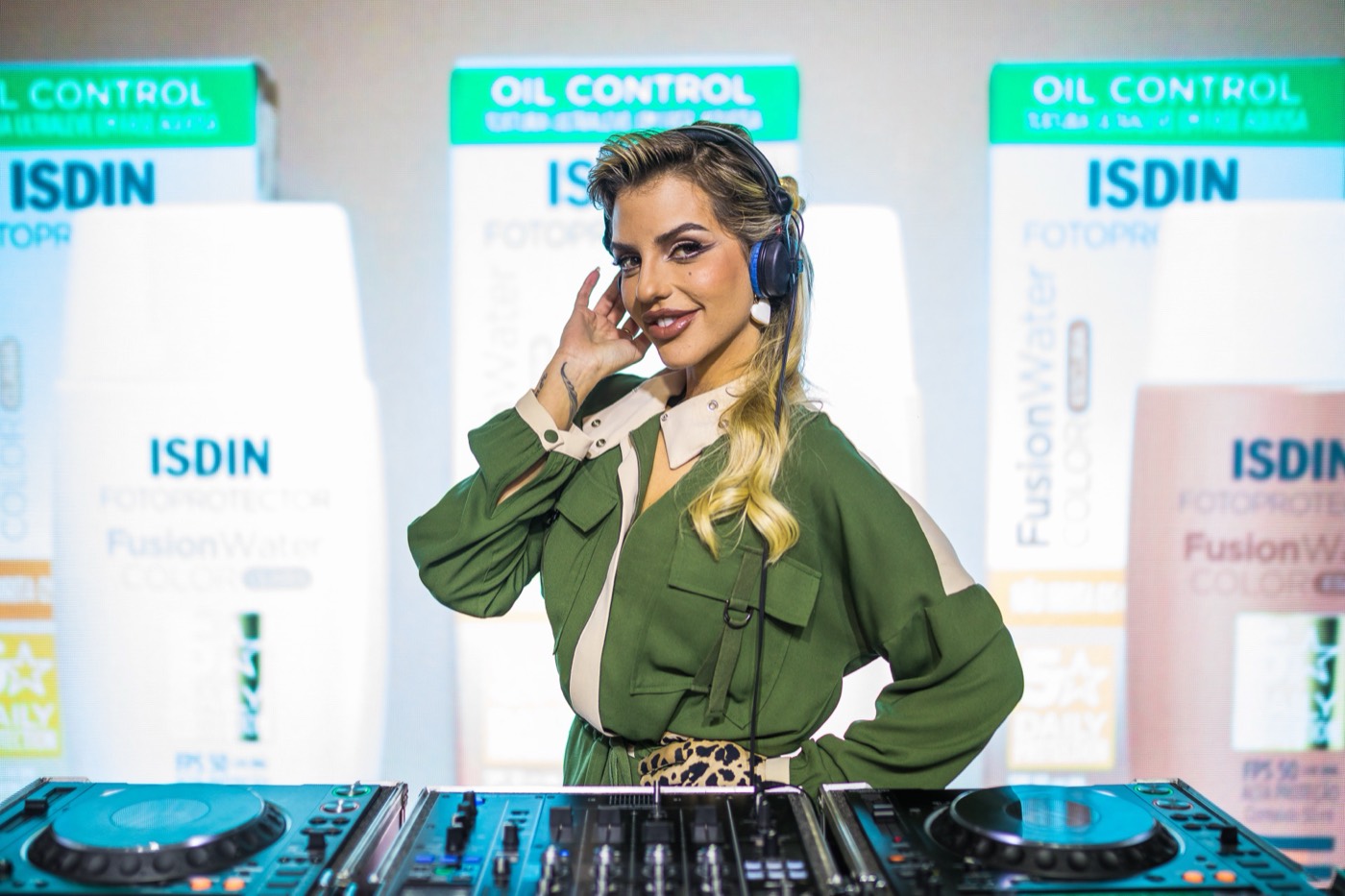 Person wearing headphones poses behind a DJ deck, in front of a screen showing "ISDIN" packaging