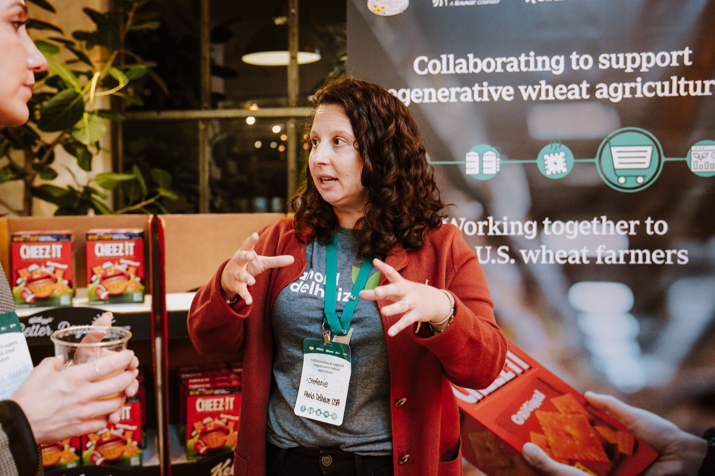Person in a red jacket and badge gestures while talking, in front of a banner and "CHEEZ-IT" boxes