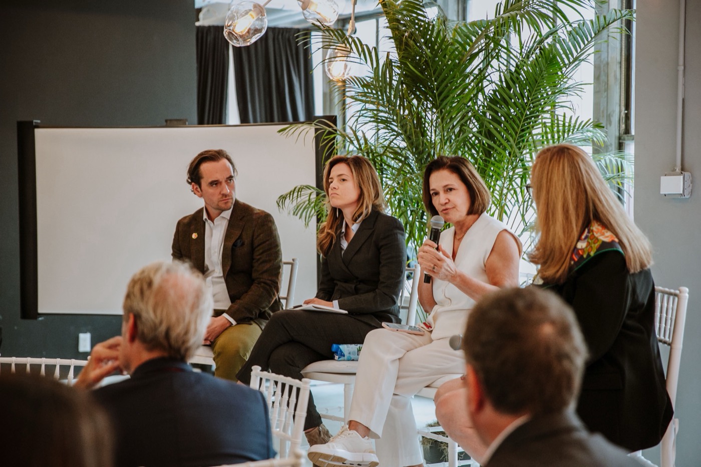 Four panelists seated before a screen and palm plant, one in white speaking into a microphone to the audience