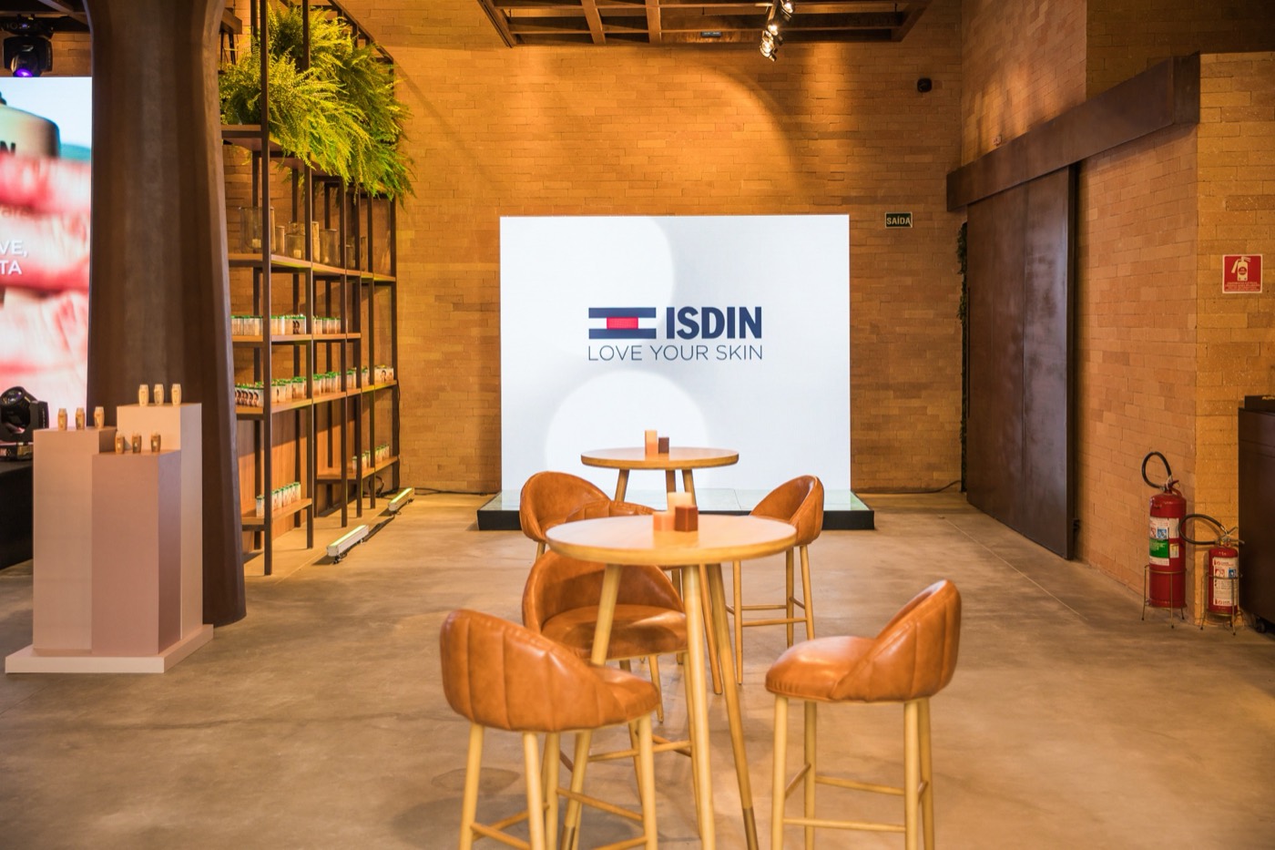 White "ISDIN LOVE YOUR SKIN" panel in a brick-walled space, with high tables and leather stools in front