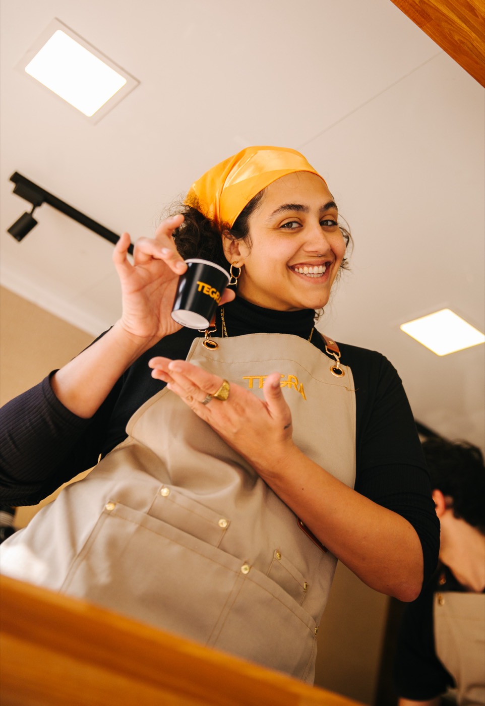 Person in a beige apron and yellow headscarf smiles holding a black cup reading "TEGRA"